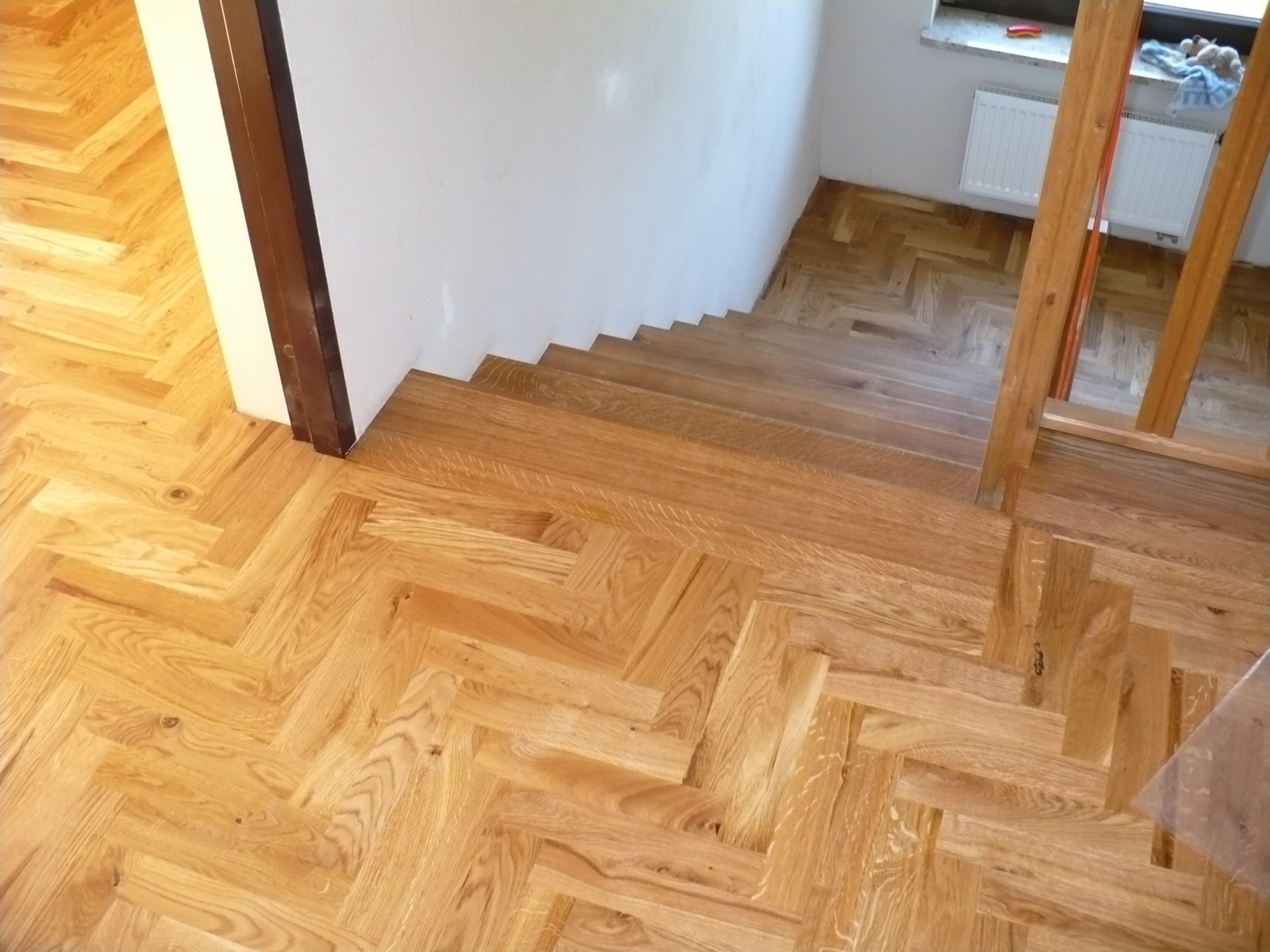 Parkiet ułożony w jodełkę na podłodze i schodach, jasne drewno, widok z góry, fragment ściany i okna w tle. Naturalne oświetlenie podkreśla strukturę drewna.