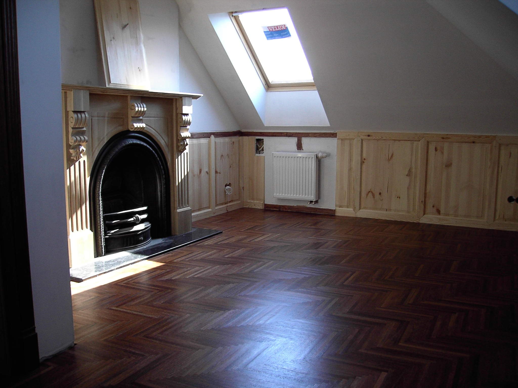 Parkiet w jodełkę, po renowacji, w jasnym pokoju na poddaszu z kominkiem i oknem dachowym. Widoczne detale wykończenia ścian i podłogi.