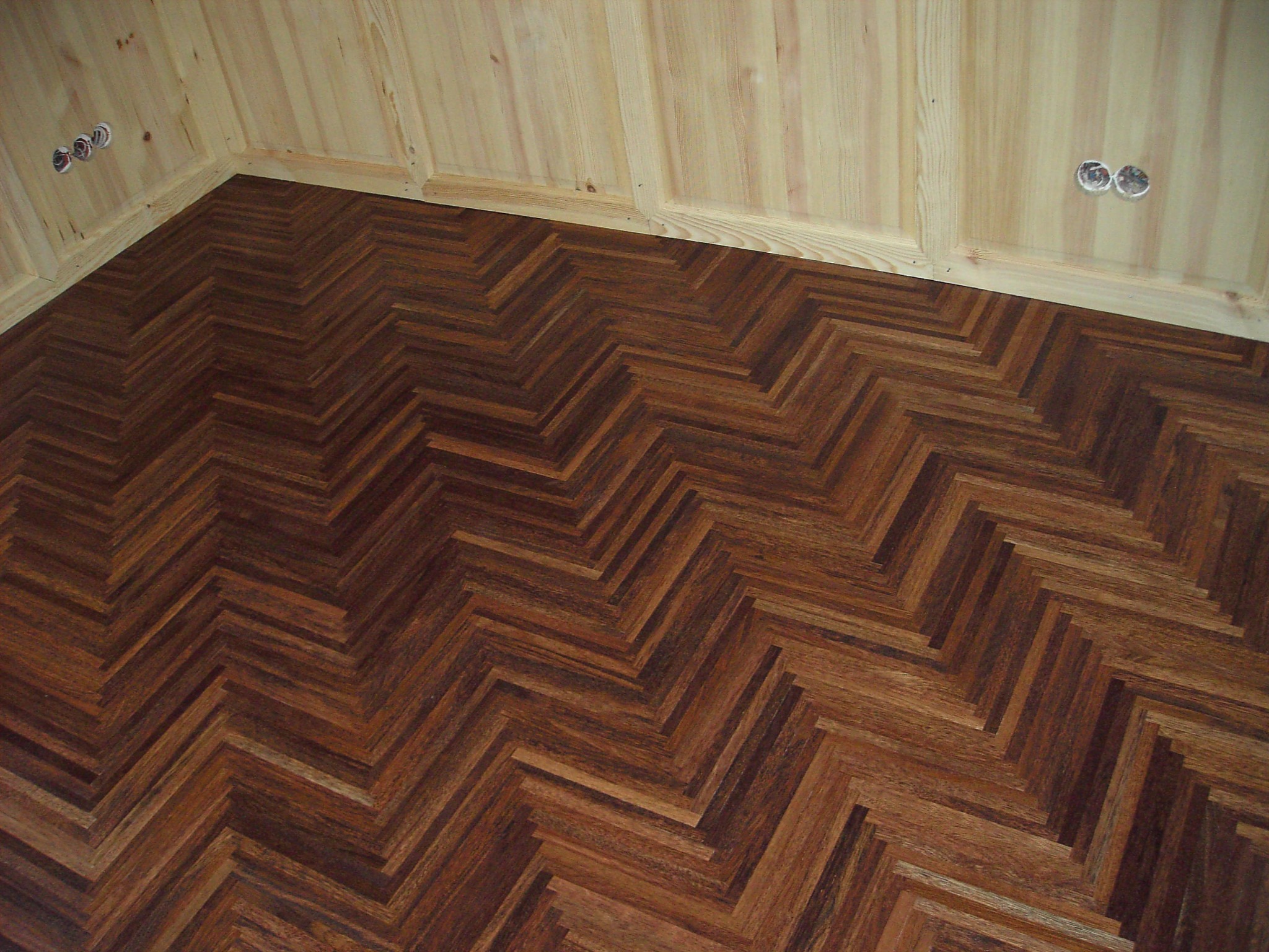 Ukończona podłoga z ciemnego parkietu ułożonego we wzór jodełki, widoczna przy jasnej, drewnianej ścianie z gniazdkami elektrycznymi.