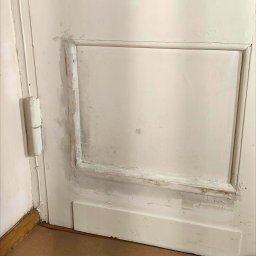 Perfect Walls Adam Andruch - Fragment białych, drewnianych drzwi z widocznymi śladami starej farby i świeżo nałożonej szpachli. Przygotowanie do malowania, widoczne zawiasy i zabezpieczona podłoga.