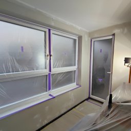 Perfect Walls Adam Andruch - Pokój przygotowany do malowania: okna i drzwi zabezpieczone folią i taśmą malarską, ściany z widocznymi śladami szpachlowania, lampa oświetlająca.