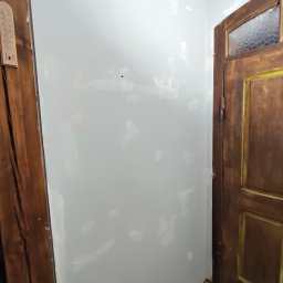 Perfect Walls Adam Andruch - Ściana po szpachlowaniu, widoczne białe plamy szpachli, przygotowana do malowania. Obok drewniana futryna drzwi i termometr na ścianie.