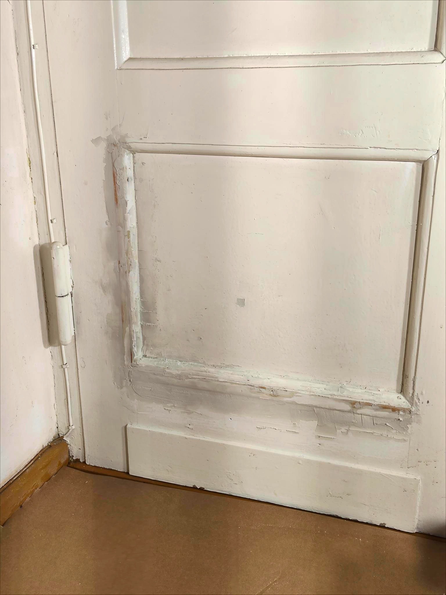 Fragment białych, drewnianych drzwi z widocznymi śladami starej farby i świeżo nałożonej szpachli. Przygotowanie do malowania, widoczne zawiasy i zabezpieczona podłoga.