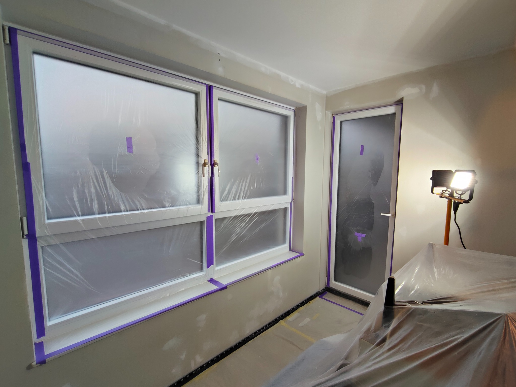 Pokój przygotowany do malowania: okna i drzwi zabezpieczone folią i taśmą malarską, ściany z widocznymi śladami szpachlowania, lampa oświetlająca.