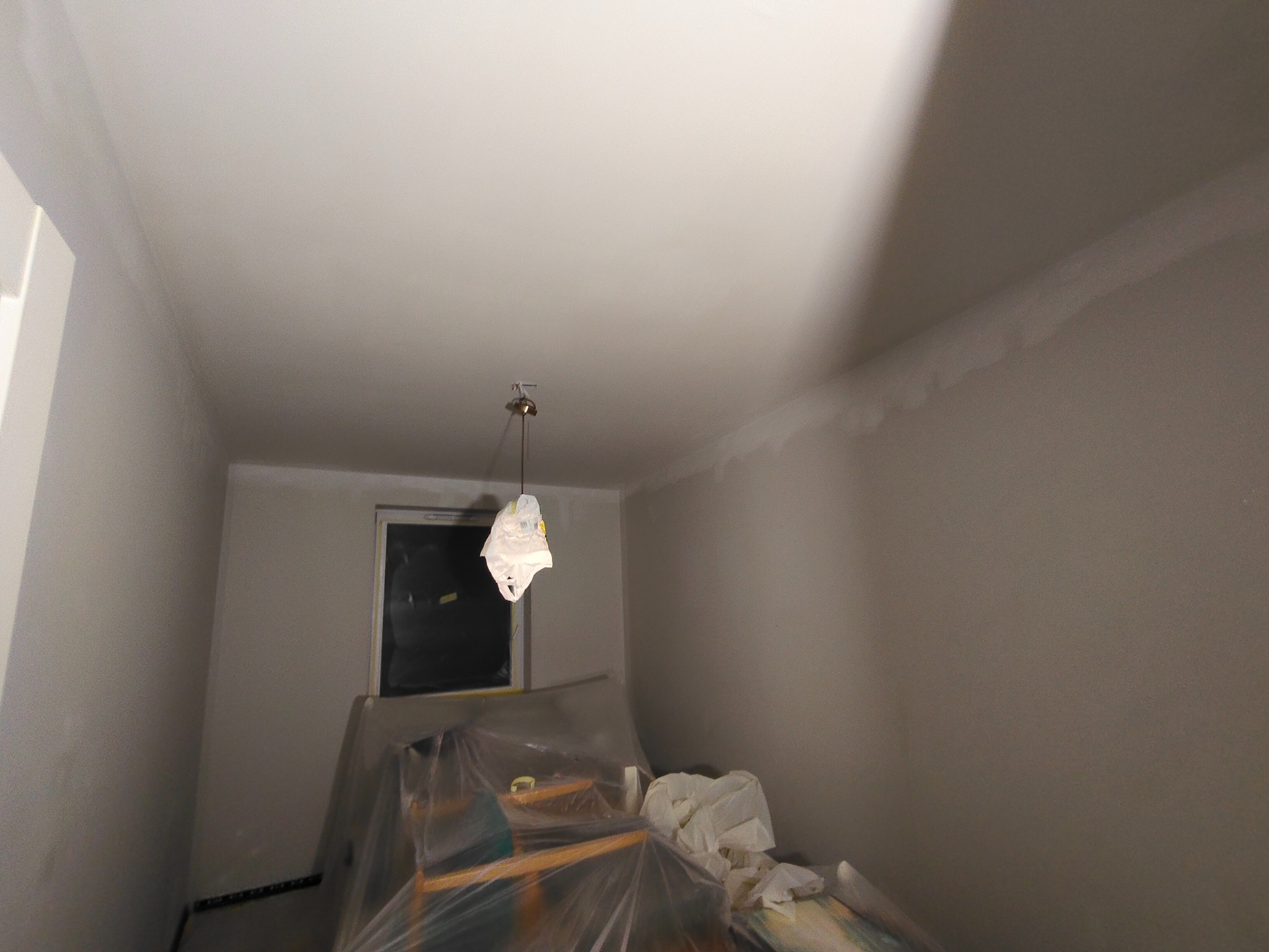 Pokój w trakcie przygotowań do malowania: zabezpieczone okno i meble folią, widoczne ślady szpachlowania na ścianach i suficie, lampa zabezpieczona workiem.