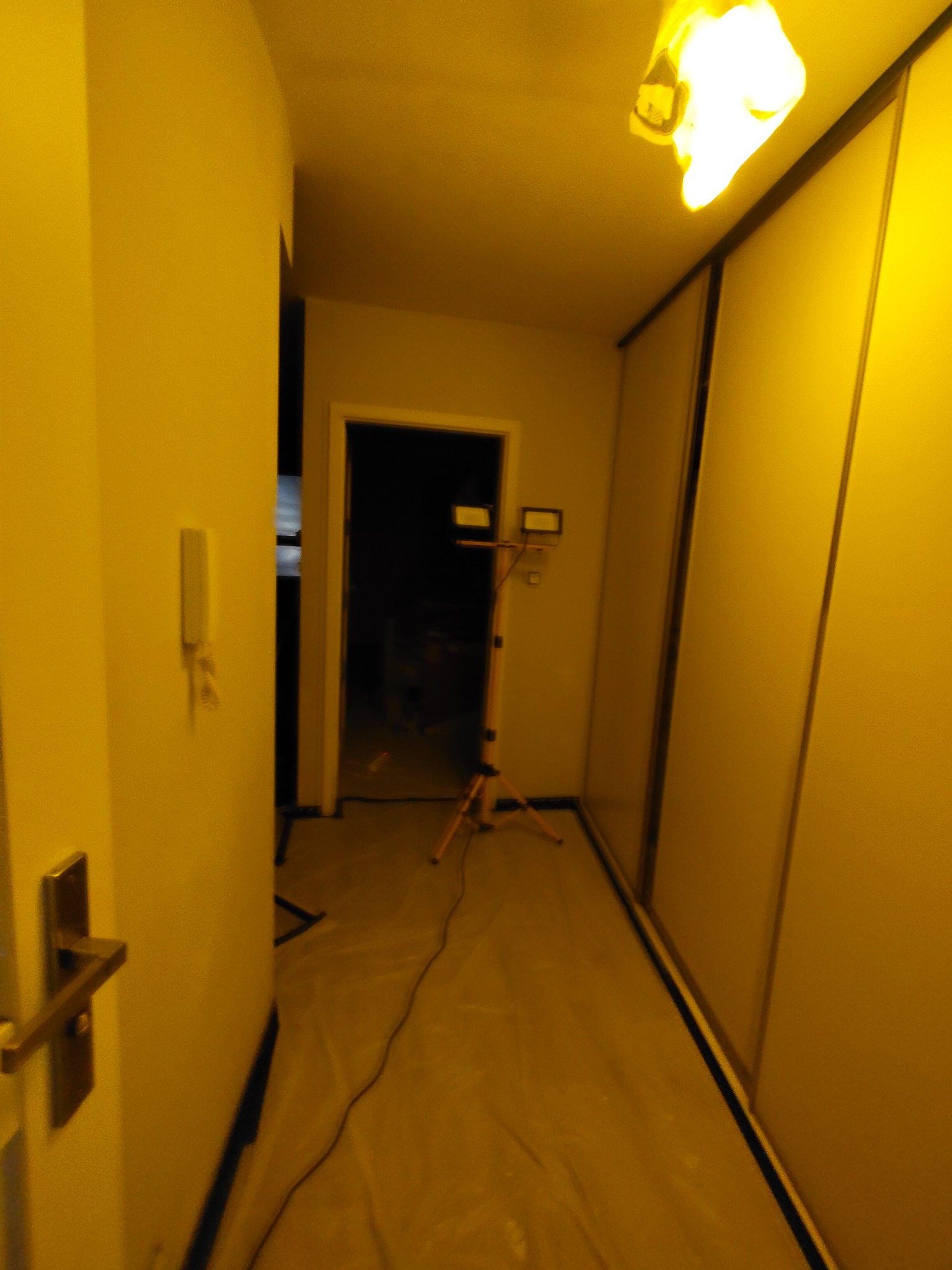 Korytarz w trakcie prac przygotowawczych: podłoga zabezpieczona folią, widoczny statyw oświetleniowy. Ściany przygotowane do malowania.