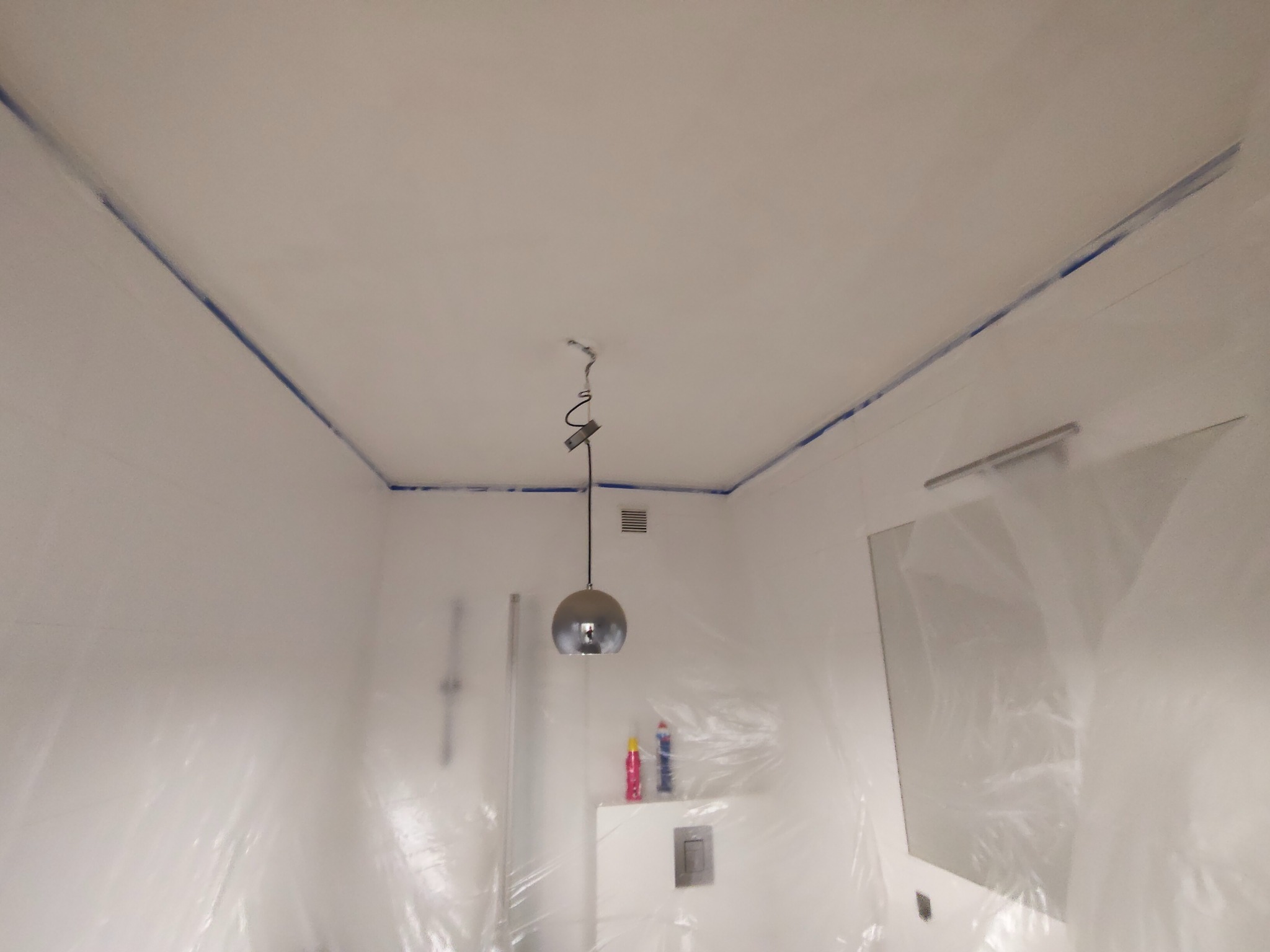 Łazienka zabezpieczona folią malarską i taśmą przed malowaniem. Widoczna lampa wisząca, fragment lustra i armatury. Przygotowanie do malowania ścian i sufitu.