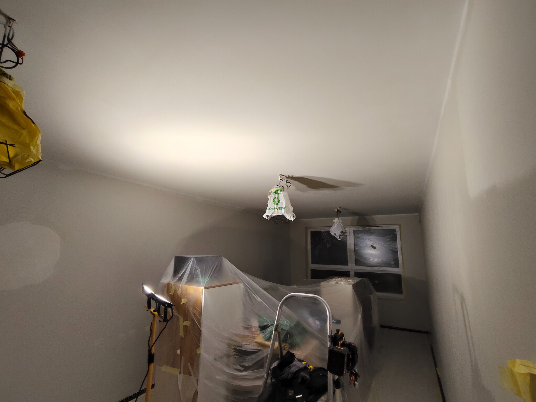 Pokój w trakcie przygotowań do malowania: zabezpieczone meble folią, widoczne ślady szpachlowania na ścianach, drabina i lampa budowlana. Przygotowanie do prac wykończeniowych.