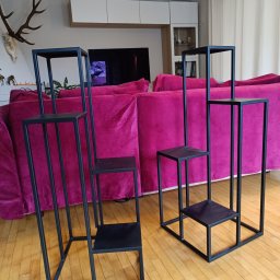 RR Desing Garage Maciej Miśkiewicz - Dwie, czarne, metalowe konstrukcje schodkowe, prawdopodobnie kwietniki, na drewnianej podłodze, z fuksjową kanapą w tle. Minimalistyczny design.