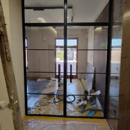 RR Desing Garage Maciej Miśkiewicz - Nowoczesna, przeszklona konstrukcja z czarnymi szprosami, prawdopodobnie w trakcie montażu. Widoczne zabezpieczenia podłogi i narzędzia.