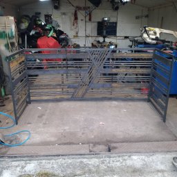 RR Desing Garage Maciej Miśkiewicz - Surowa, metalowa brama wjazdowa z ukośnymi zdobieniami, w trakcie produkcji w warsztacie. Widoczne narzędzia i elementy wyposażenia. Szare tło, betonowa podłoga.