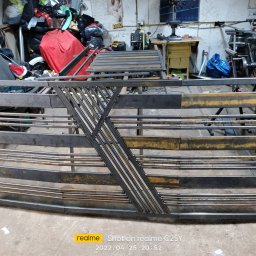 RR Desing Garage Maciej Miśkiewicz - Surowa, spawana brama z metalowymi prętami i ukośnymi wzmocnieniami, widoczna w warsztacie. Elementy konstrukcyjne w trakcie realizacji, przed malowaniem.