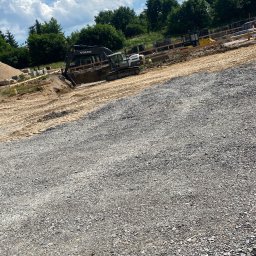 Usługi Remontowo Budowlane Dawid Szczepaniak - Praca ziemna: koparka Volvo na placu budowy, przygotowuje teren pod fundamenty. Widoczne szalunki i usypany żwir. Błękitne niebo z chmurami w tle.