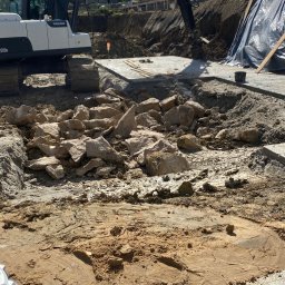 Usługi Remontowo Budowlane Dawid Szczepaniak - Prace ziemne: koparka Volvo na placu budowy, wykop z kamieniami i piaskiem, widoczne fundamenty i zabezpieczenia wykopu. Słoneczny dzień.