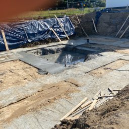 Usługi Remontowo Budowlane Dawid Szczepaniak - Wykop pod fundamenty z częściowo wylanym betonem, zabezpieczony folią i drewnianymi podporami. Widoczne ślady ziemi i piasku na betonie.