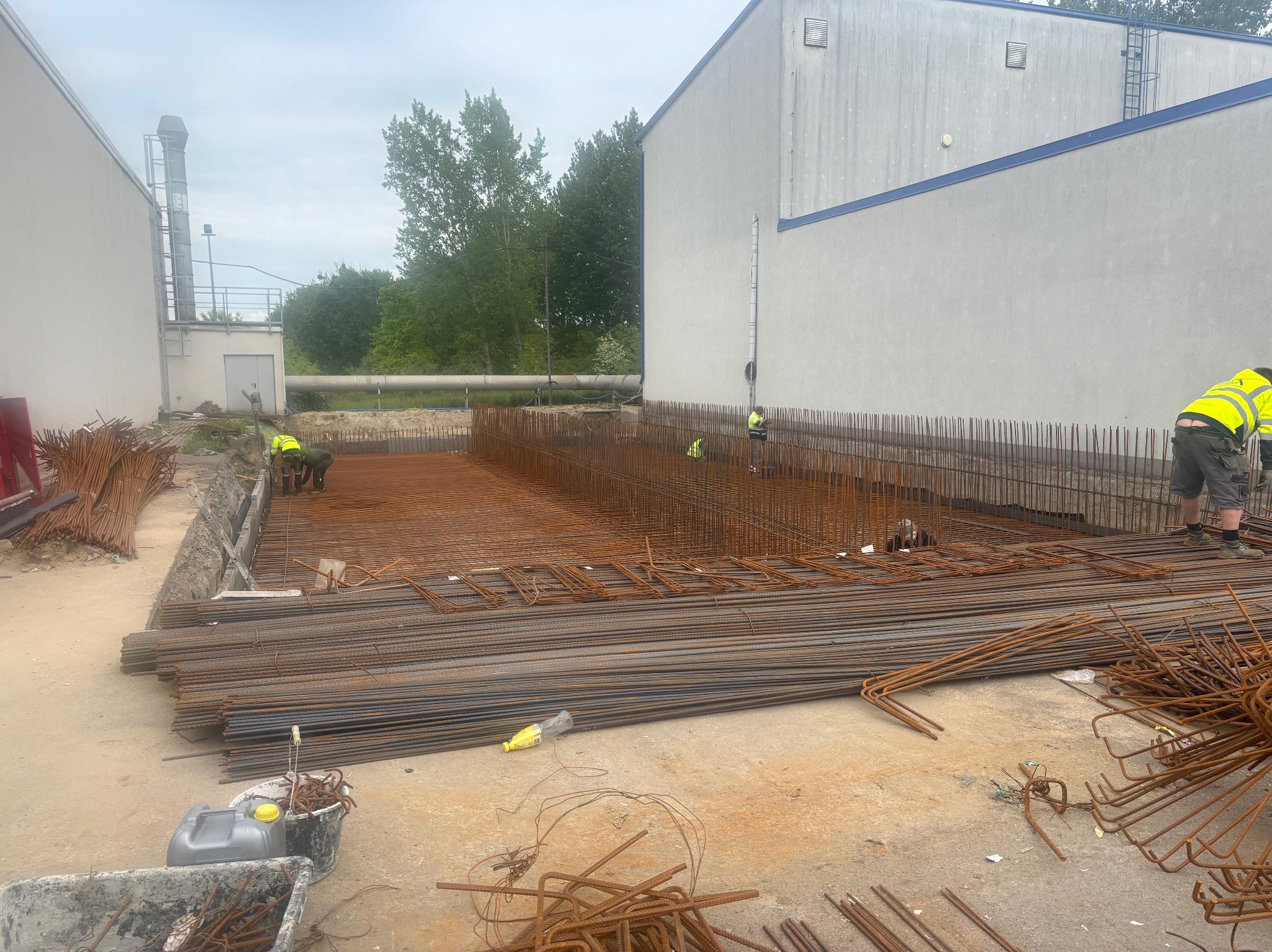 Zbrojenie fundamentów budynku przemysłowego z widocznymi pracownikami w kamizelkach odblaskowych. Stalowe pręty ułożone w siatkę, przygotowane do zalania betonem.