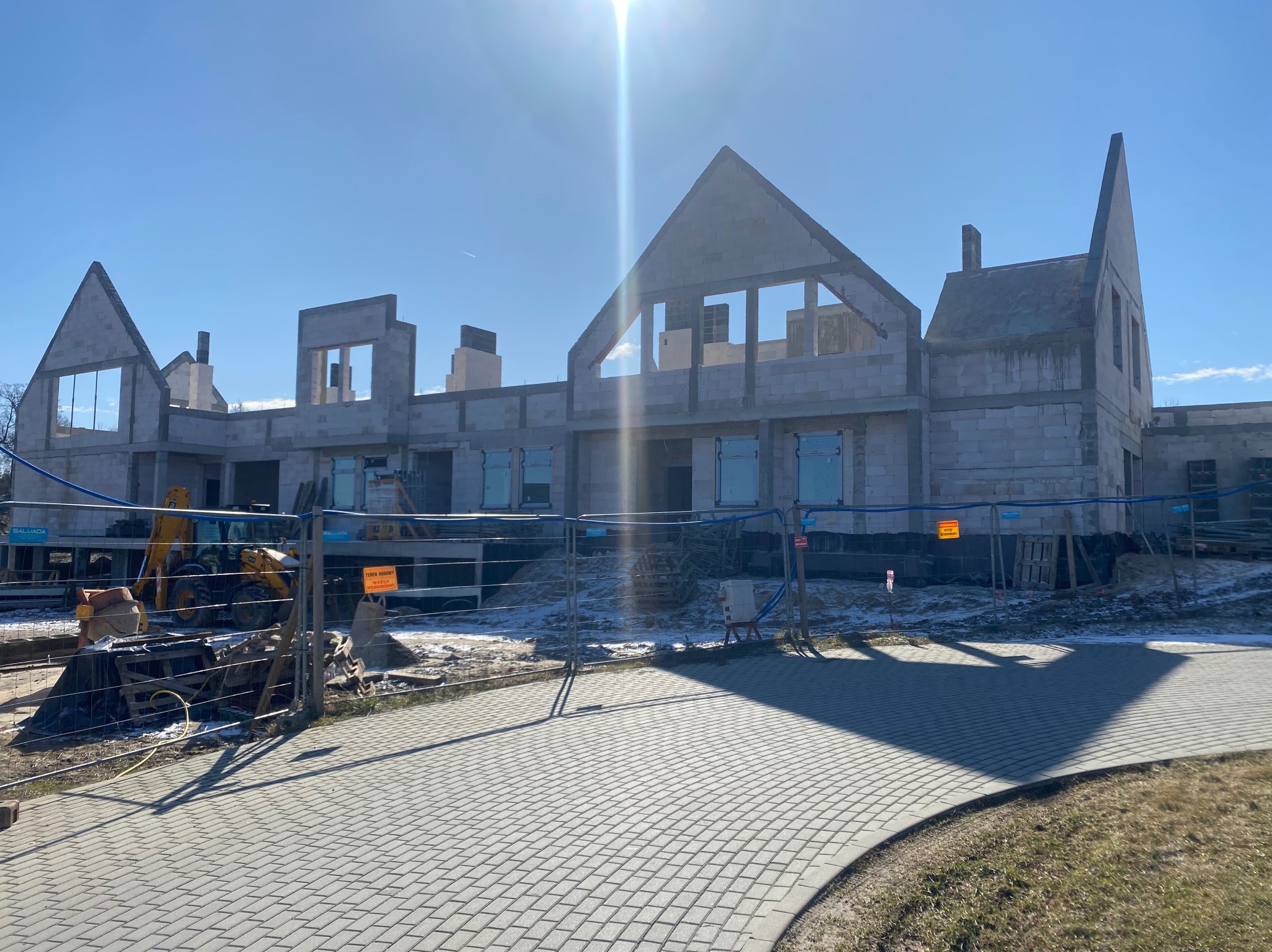 Surowy stan budynku z betonu komórkowego w trakcie budowy, widoczny zza ogrodzenia placu budowy w słoneczny dzień. Częściowo ośnieżony teren.