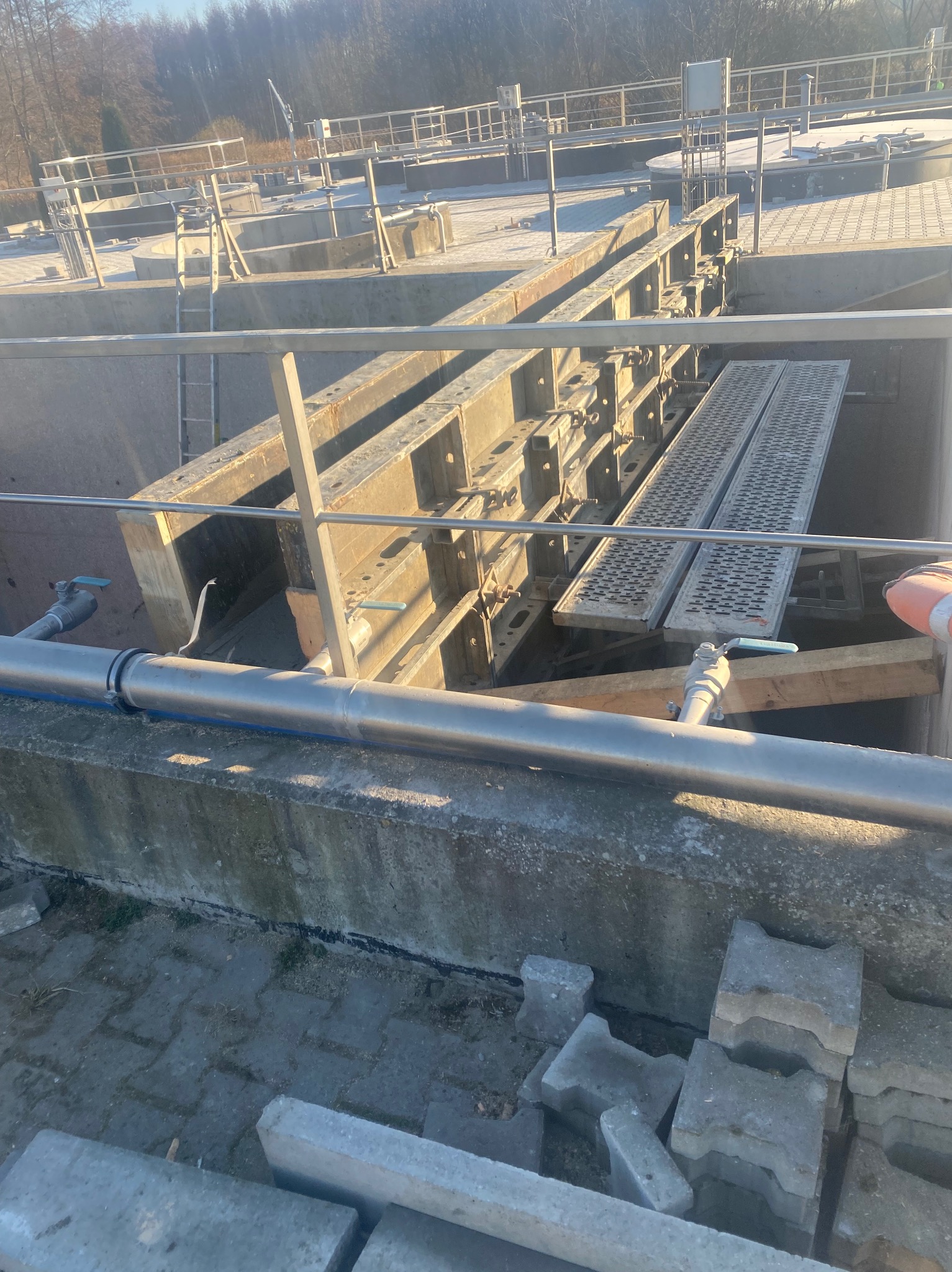 Szare, betonowe elementy konstrukcyjne w trakcie budowy, widoczne metalowe szalunki i rury. Elementy ułożone na placu budowy, w tle drzewa.