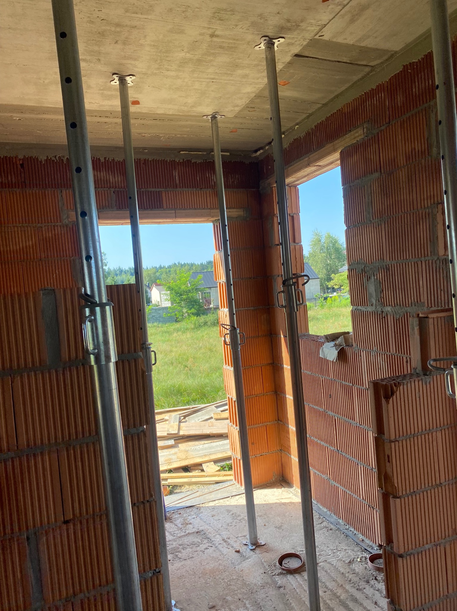 Wnętrze w trakcie budowy z pustaków ceramicznych, wsparte metalowymi stemplami. Widok na zewnątrz przez otwory okienne, w tle zieleń i zabudowa jednorodzinna.