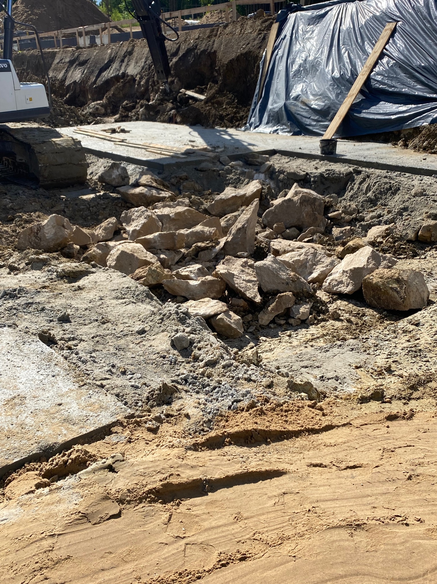 Fundamenty w trakcie budowy: wykop, częściowo zalany beton, kamienie i ciężki sprzęt budowlany. Widoczne zabezpieczenia wykopu.