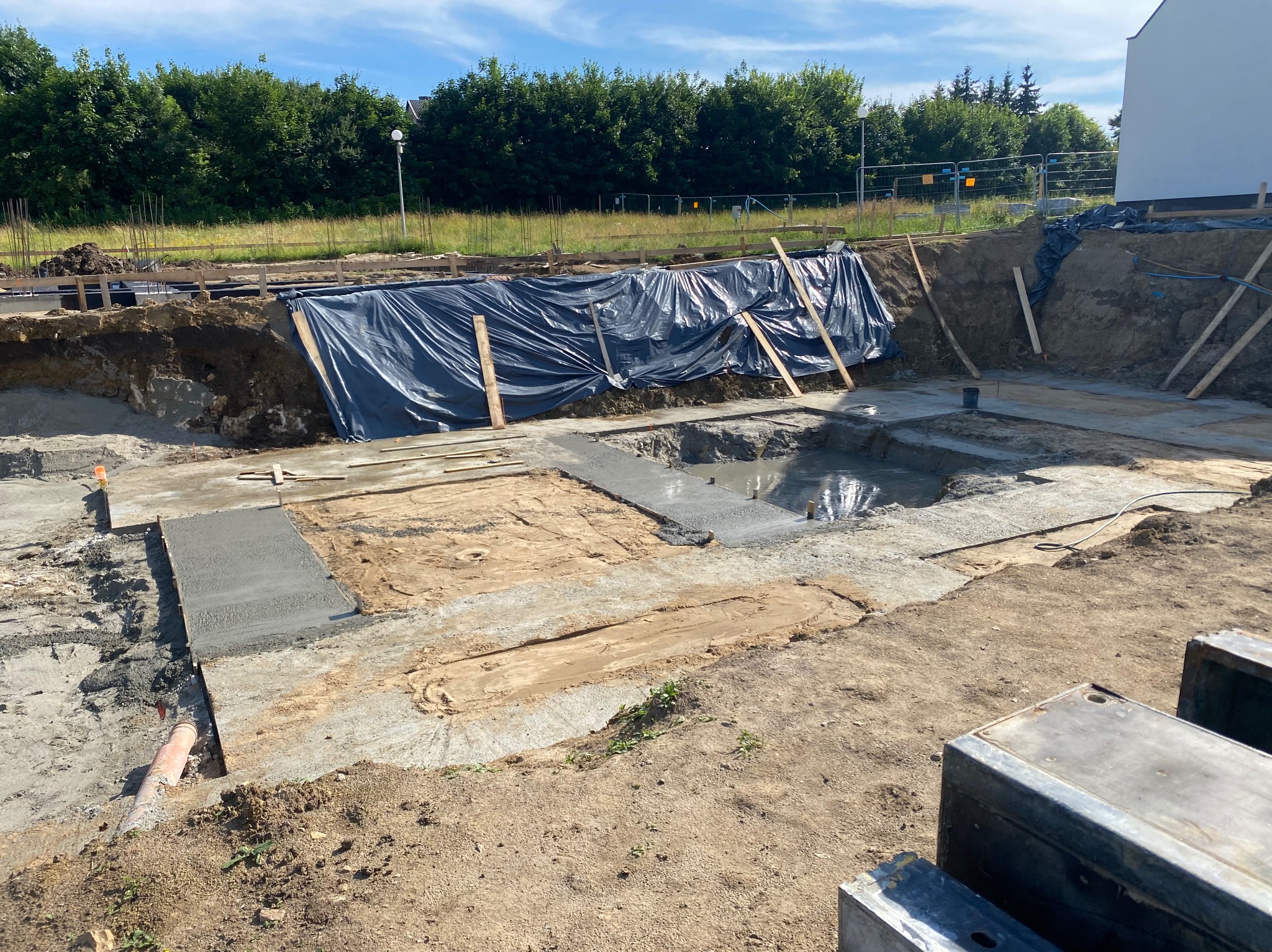Wykop fundamentów z częściowo wylanym betonem, zabezpieczony folią i drewnianymi podporami. Widoczne elementy szalunków i rury. W tle zieleń i fragment budynku.