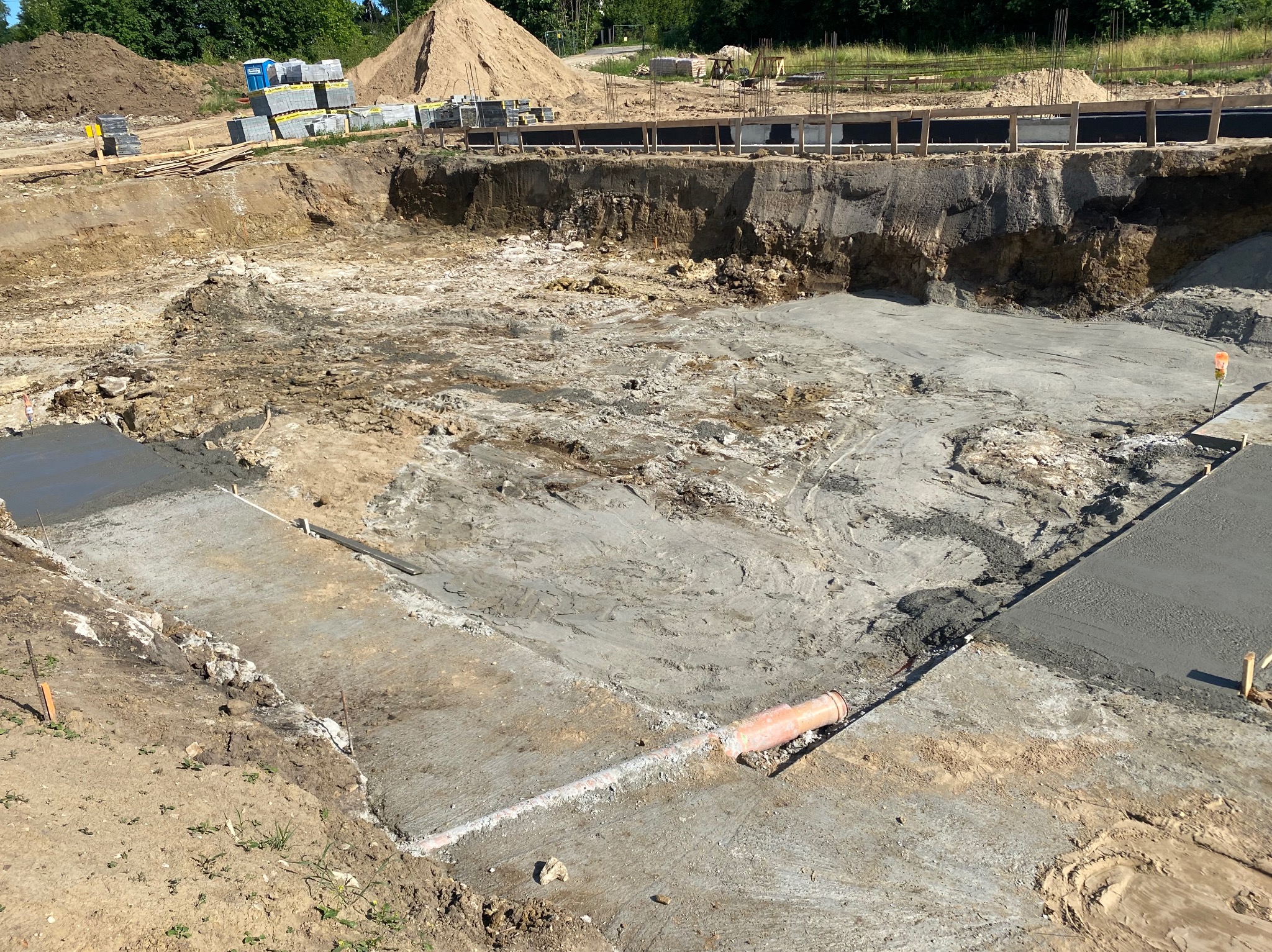 Wykop fundamentowy z częściowo wylanym betonem, widoczne rury i przygotowane szalunki. Na górze hałda ziemi i materiały budowlane.