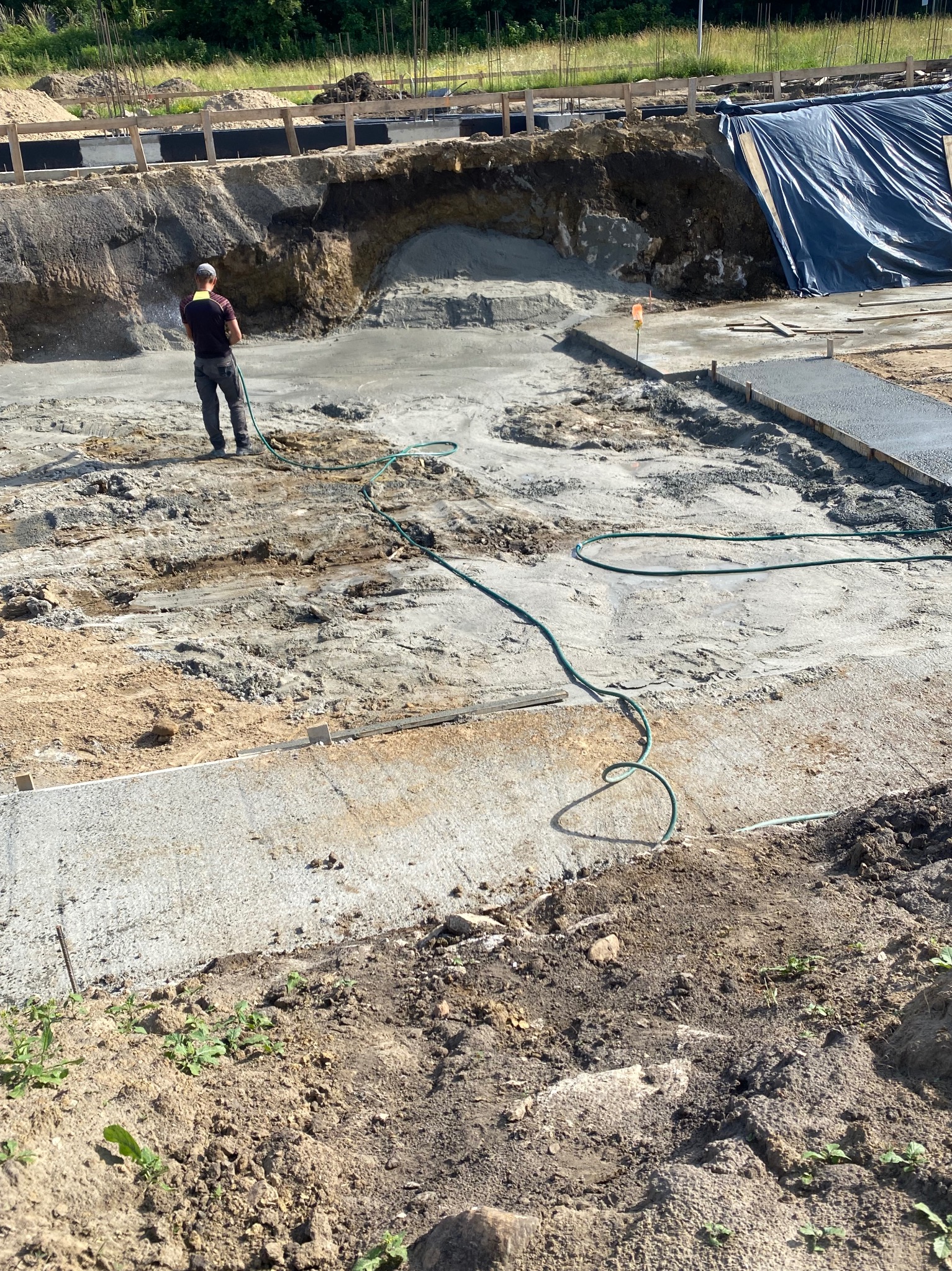 Wylewka fundamentów w trakcie prac. Robotnik podlewa beton w wykopie, widoczne szalunki i zabezpieczenie wykopu folią. Prace ziemne w toku.