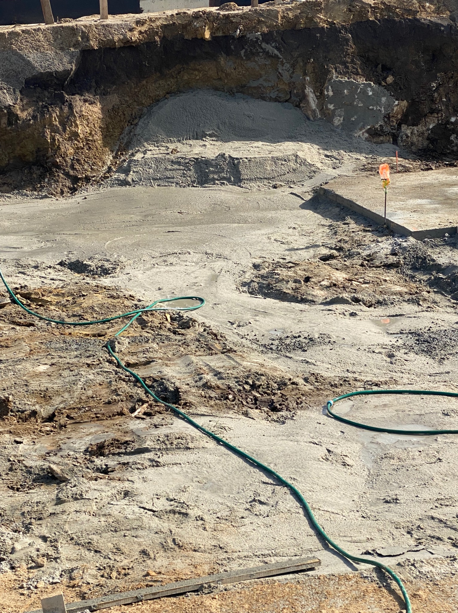 Wykonane wykopy pod fundamenty z widocznymi warstwami gruntu i częściowo wylanym betonem. W tle zabezpieczona skarpa. Wąż ogrodowy na pierwszym planie.
