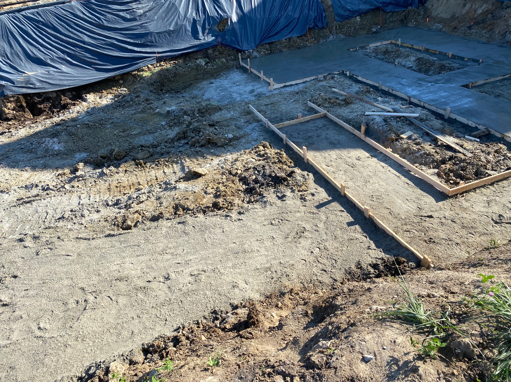 Wykop pod fundamenty z częściowo wylanym betonem i szalunkami. Widoczna czarna folia ochronna. Ujęcie z góry, widać ziemię i roślinność.