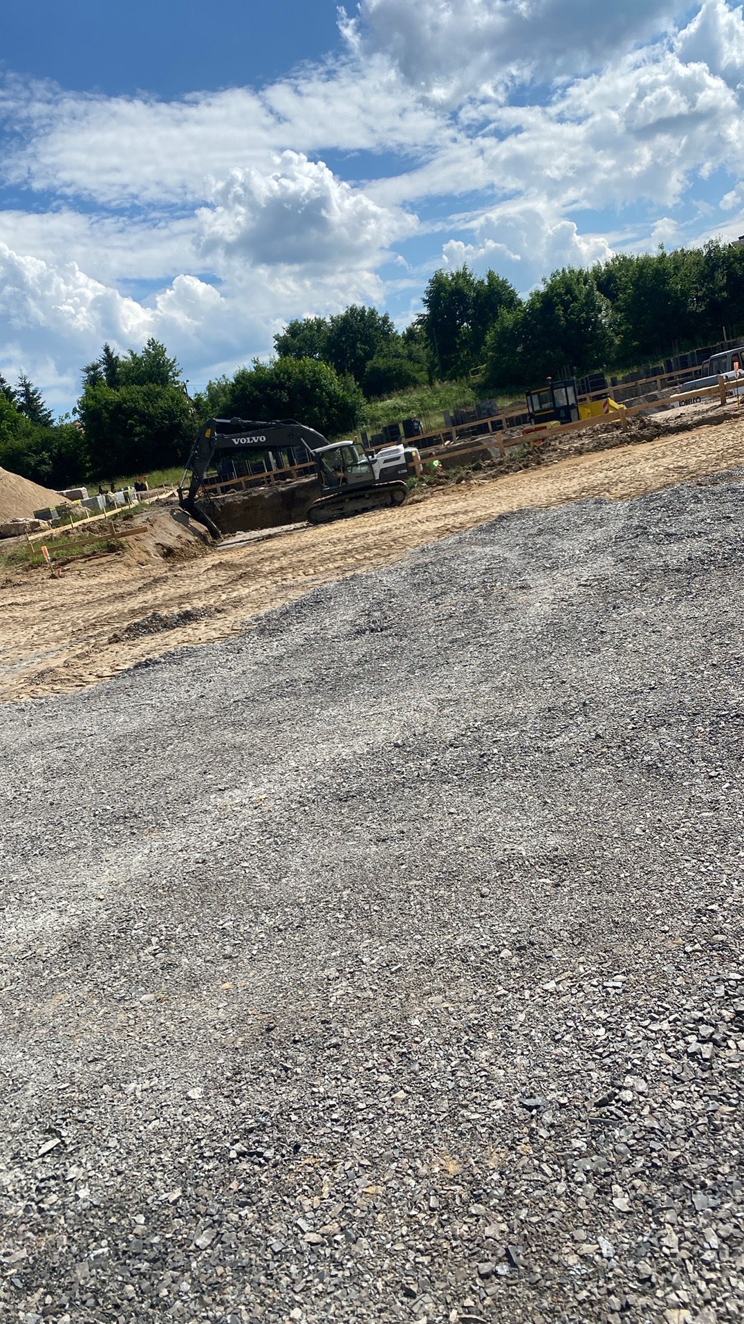 Praca ziemna: Koparka Volvo na placu budowy, przygotowuje teren pod fundamenty. Widoczne szalunki i materiały budowlane. Słoneczny dzień, błękitne niebo z chmurami.