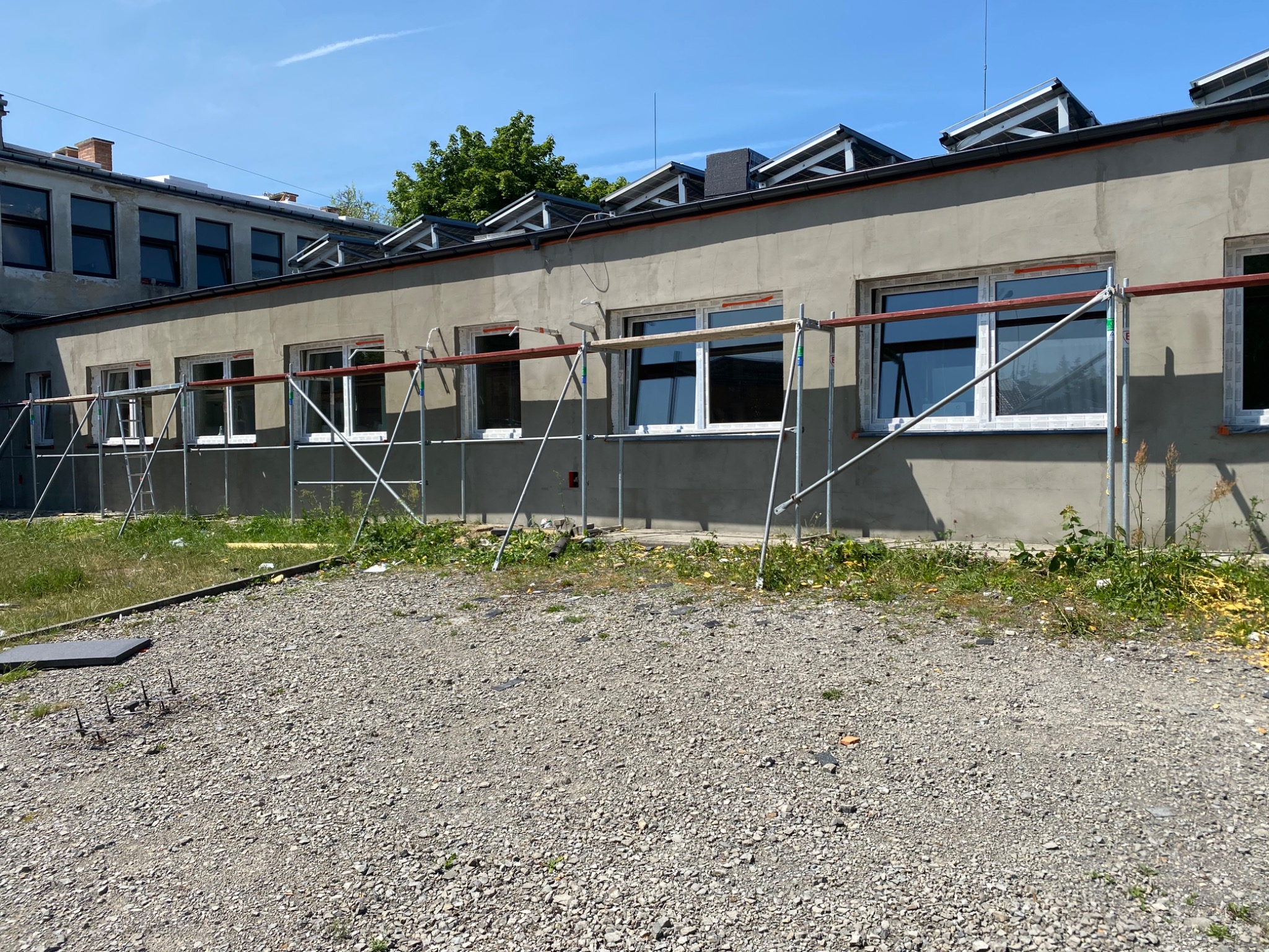 Szary budynek w trakcie prac wykończeniowych, z oknami i rusztowaniami na zewnątrz. Widoczne fragmenty elewacji i otoczenie budynku.