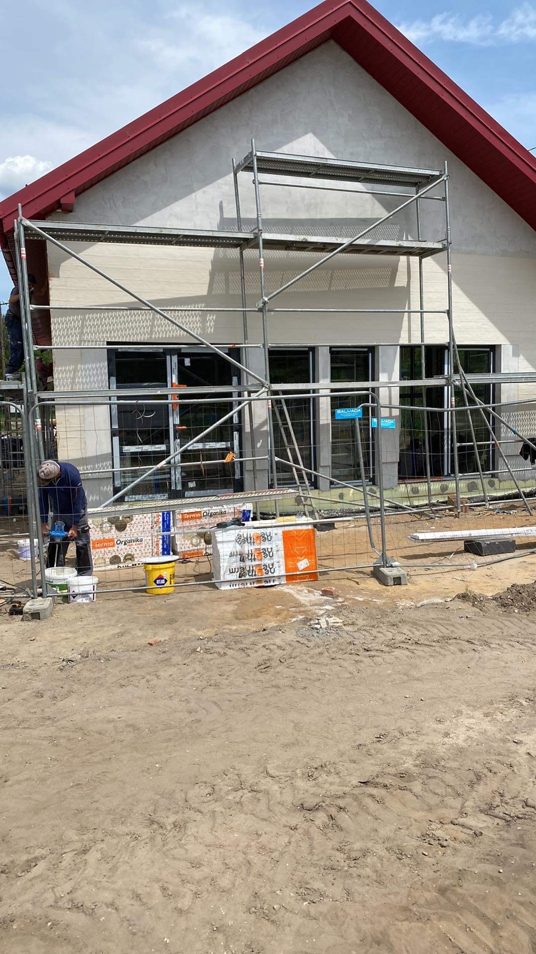 Prace wykończeniowe na elewacji domu z czerwonym dachem. Widoczne rusztowania, materiały izolacyjne i pracownik przygotowujący zaprawę.