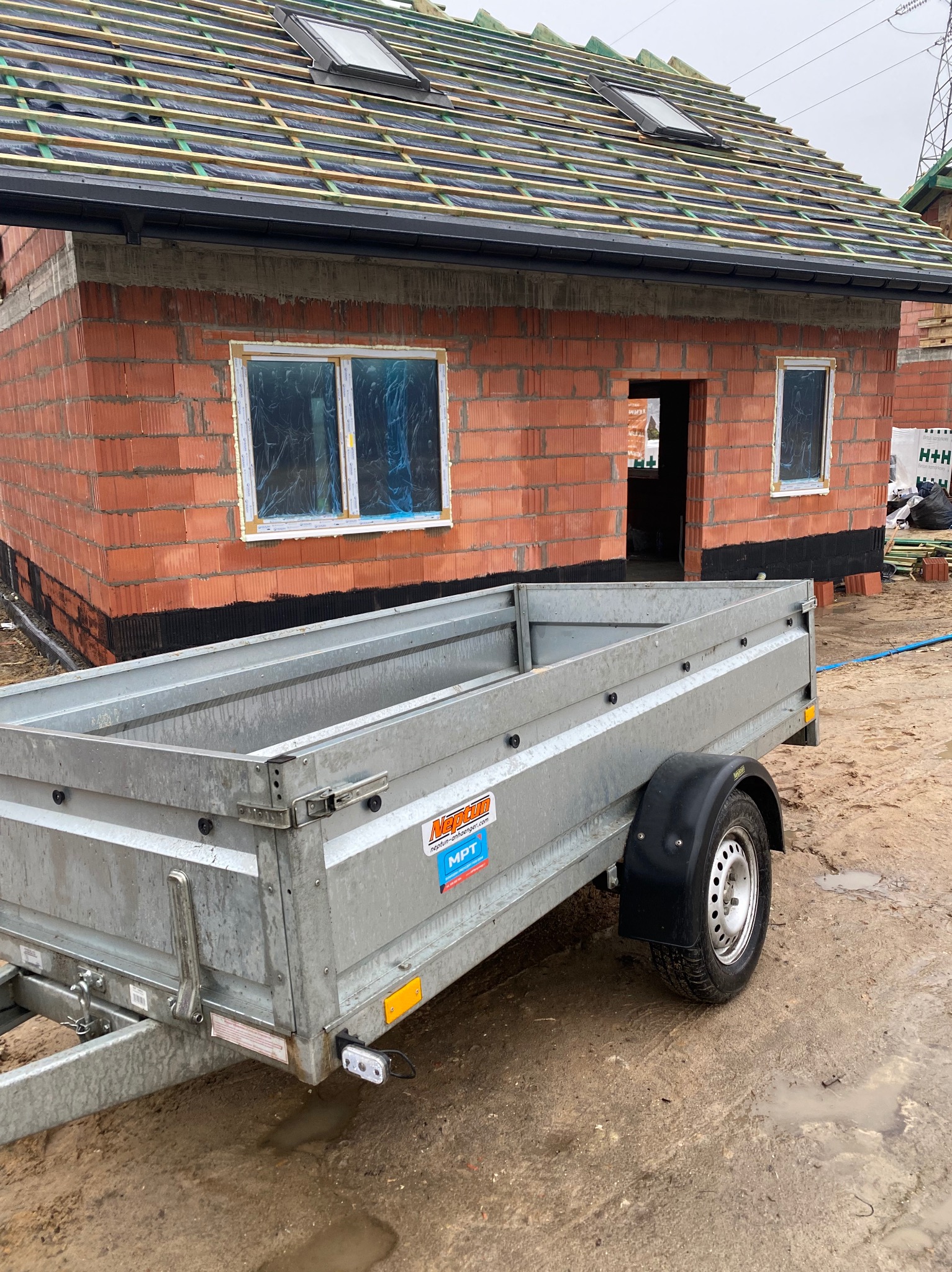 Budowa domu z czerwonej cegły w trakcie realizacji, widoczny fragment dachu z oknami, okna zabezpieczone folią, na pierwszym planie przyczepka.