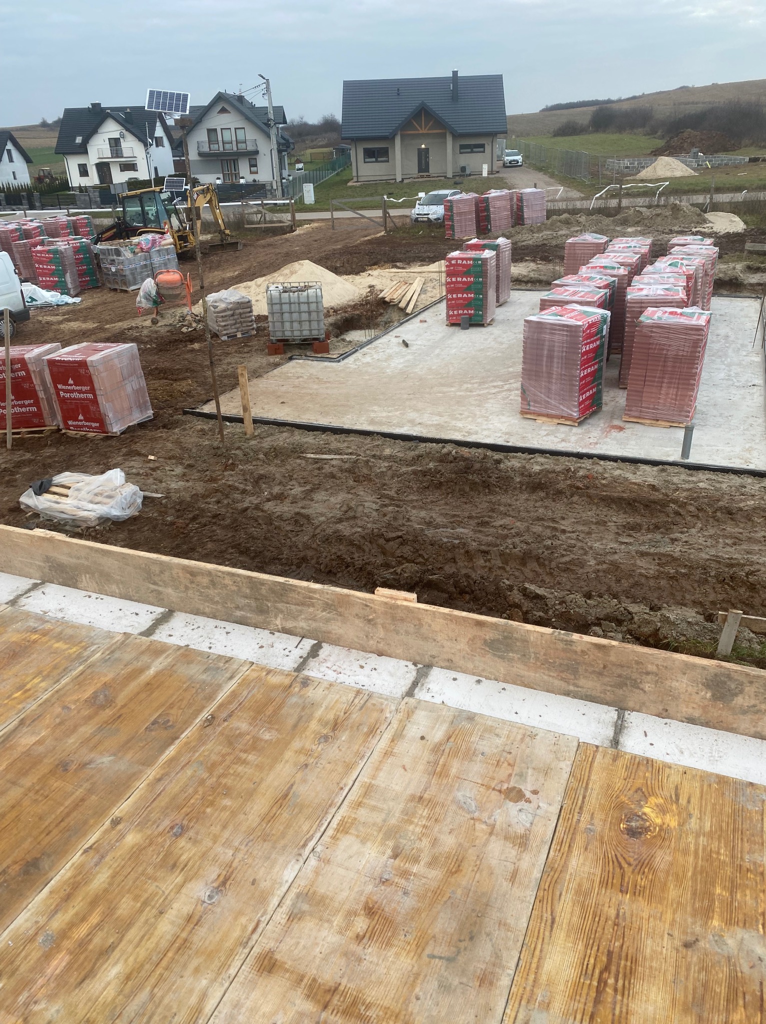 Fundamenty domu jednorodzinnego w trakcie budowy. Widoczne bloczki Porotherm, wykop, szalunki i przygotowany teren pod dalsze prace. W tle zabudowa mieszkalna.