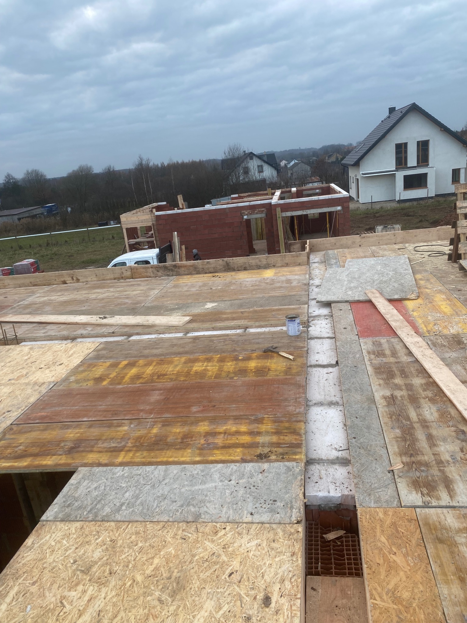 Budowa domu w trakcie: widok z góry na drewniany szalunek stropu, fragment muru z czerwonej cegły i dom w tle. Na szalunku leżą narzędzia i puszka farby.