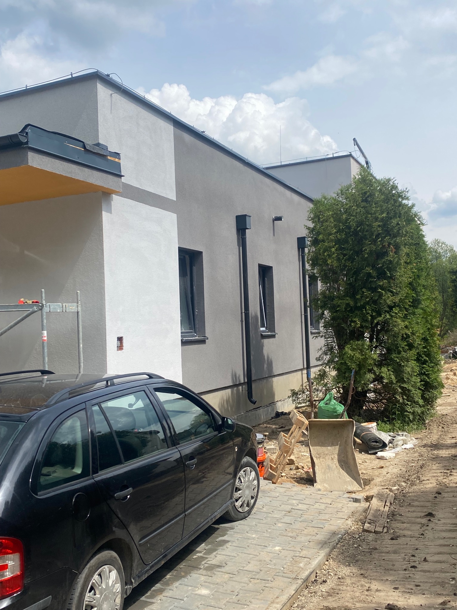 Nowoczesny dom jednorodzinny z szarą elewacją i czarnymi oknami. Na pierwszym planie zaparkowany czarny samochód i materiały budowlane.