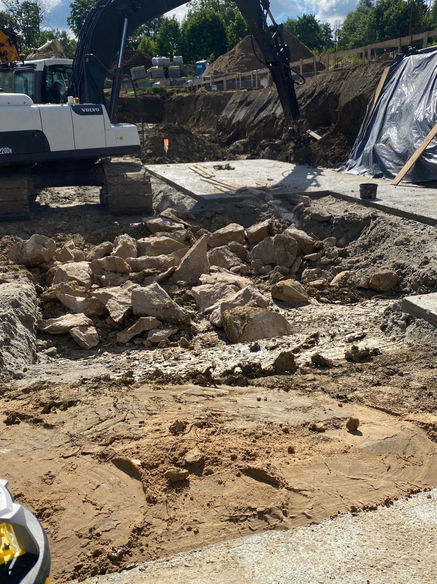 Prace ziemne: koparka Volvo na placu budowy, wykop z kamieniami i piaskiem, widoczne fundamenty i zabezpieczenia wykopu. Słoneczny dzień.