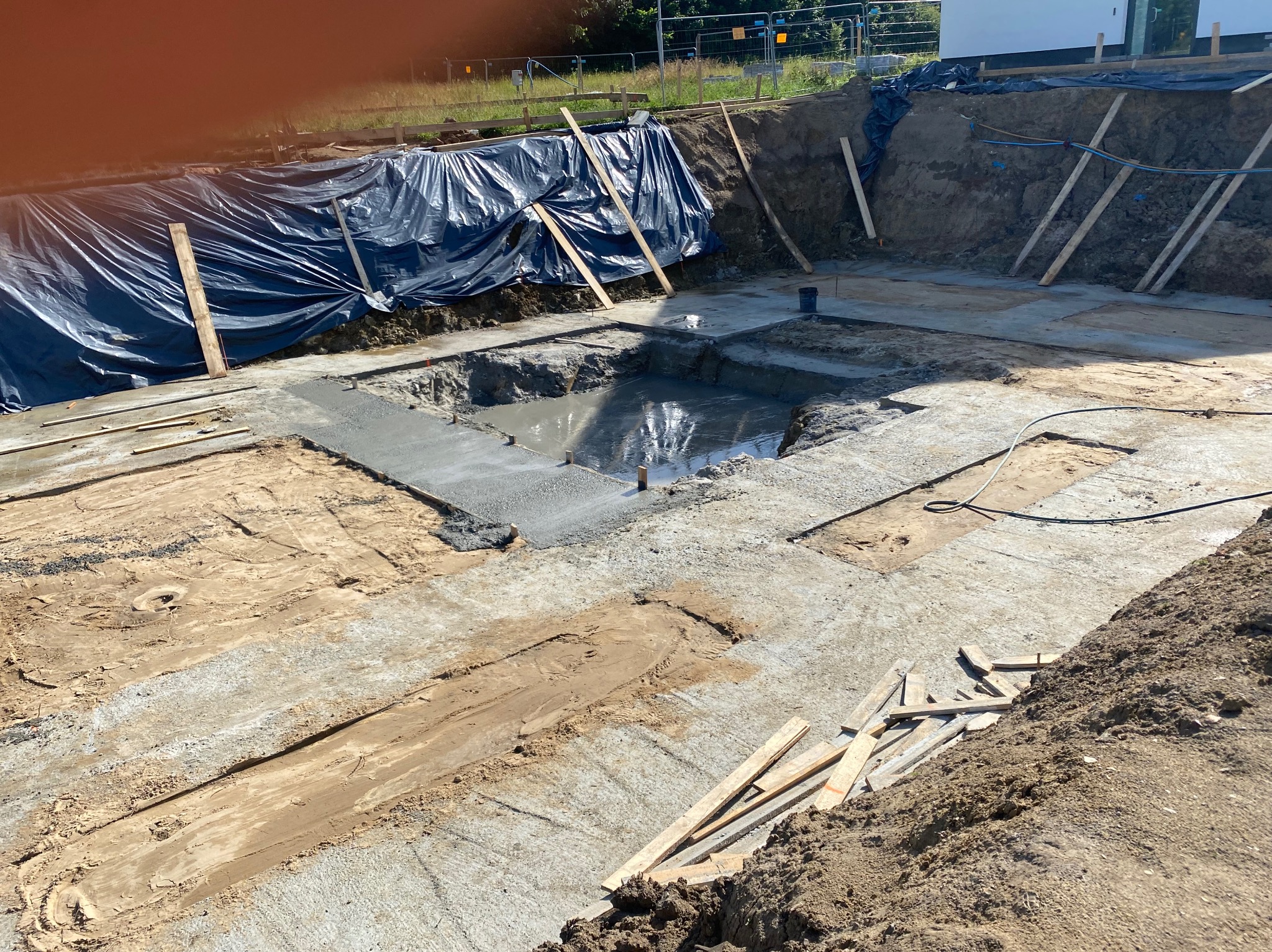Wykop pod fundamenty z częściowo wylanym betonem, zabezpieczony folią i drewnianymi podporami. Widoczne ślady ziemi i piasku na betonie.