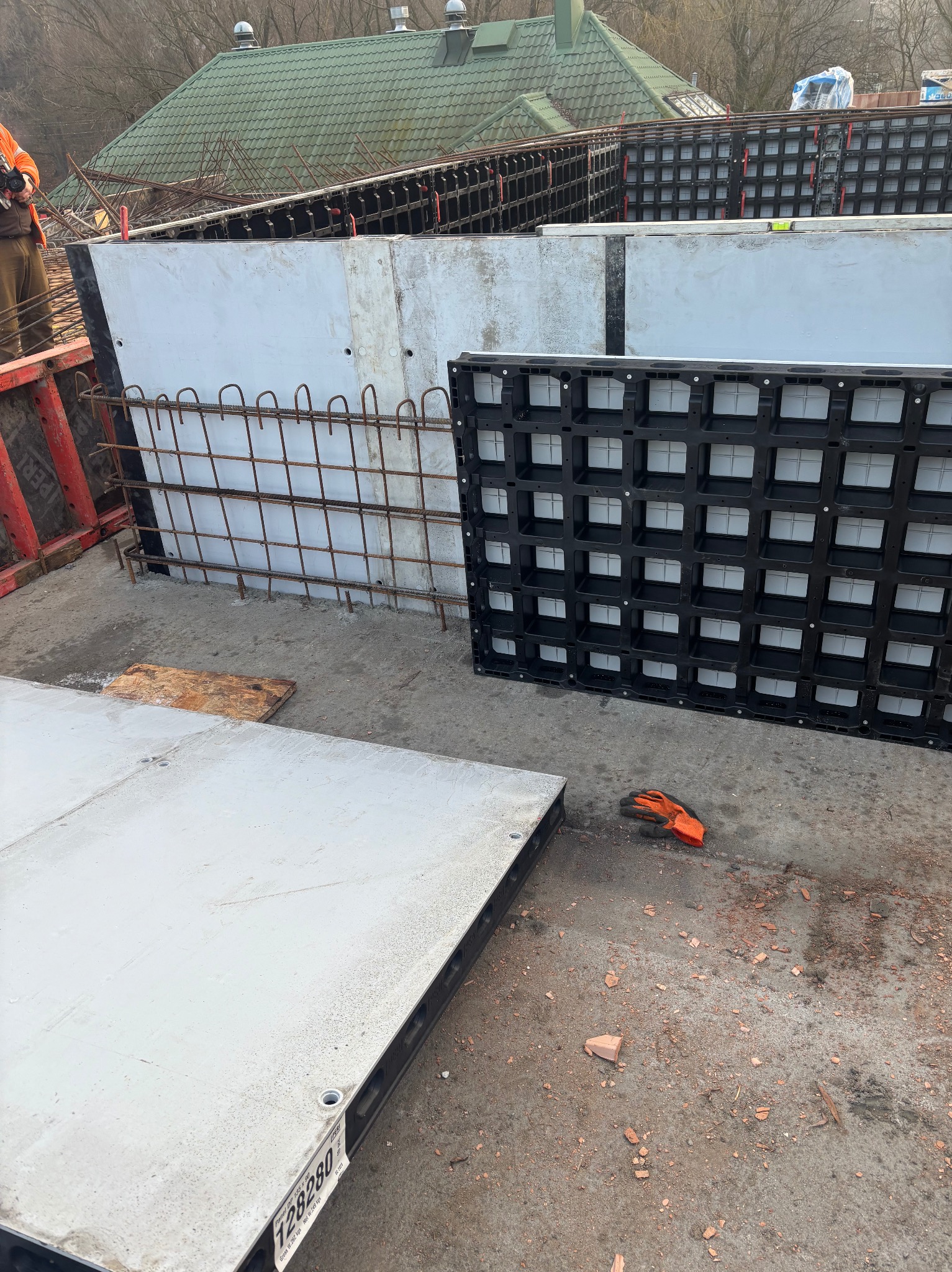 Szare szalunki zbrojeniowe, częściowo wypełnione betonem, tworzące fundament budynku. Widoczne pomarańczowe rękawice robocze i fragment dachu w tle.