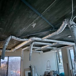 R1 Technika Łukasz Bień - Srebrne, elastyczne rury wentylacyjne zamontowane pod surowym, betonowym sufitem w jasnym pomieszczeniu z oknami. Widoczne drabiny i narzędzia.