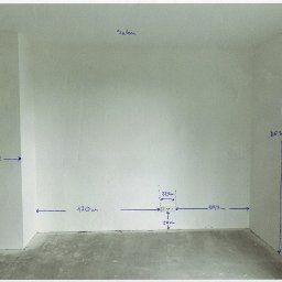 Pusty pokój w trakcie remontu z naniesionymi odręcznie wymiarami na ścianach, widoczne gniazdka elektryczne. Szary beton na podłodze, białe ściany. Salon w trakcie prac.