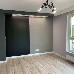 Nowoczesne wnętrze: drewnopodobne panele, ściana z szarą i czarną fakturą, designerska lampa sufitowa, okno z widokiem na zieleń. Minimalizm i elegancja.