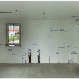 Ściana z naniesionymi odręcznymi pomiarami i oznaczeniami instalacji elektrycznej, widoczne kable. Surowy stan pomieszczenia przed pracami wykończeniowymi.