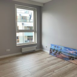 LukasFliz - Jasne panele podłogowe w pokoju z szarą ścianą, oknem z widokiem na budynek i obrazem z motywem plaży opartym o ścianę. Widoczne gniazdka elektryczne i białe listwy przypodłogowe.