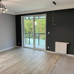 LukasFliz - Jasne panele podłogowe w nowym pokoju z balkonem i grzejnikiem na ciemnej ścianie. Widok na zieleń za oknem. Minimalistyczna lampa sufitowa.