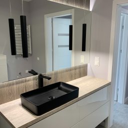 LukasFliz - Nowoczesna łazienka z czarną, prostokątną umywalką nablatową, minimalistyczną armaturą i podświetlanym lustrem. Widoczne wiszące lampy i płytki imitujące drewno.