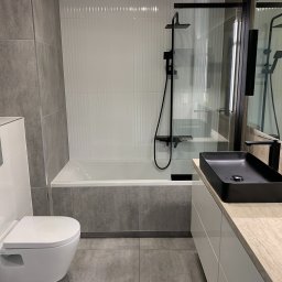 LukasFliz - Nowoczesna łazienka z wanną obudowaną szarą płytką, białą armaturą i czarną, prostokątną umywalką nablatową. Minimalistyczny design, szaro-biała kolorystyka.
