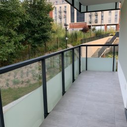 LukasFliz - Nowoczesny balkon z szarymi płytkami i szklaną balustradą. Widok na zieleń i budynek w tle. Minimalistyczny design, jasne kolory, świeżo po remoncie.