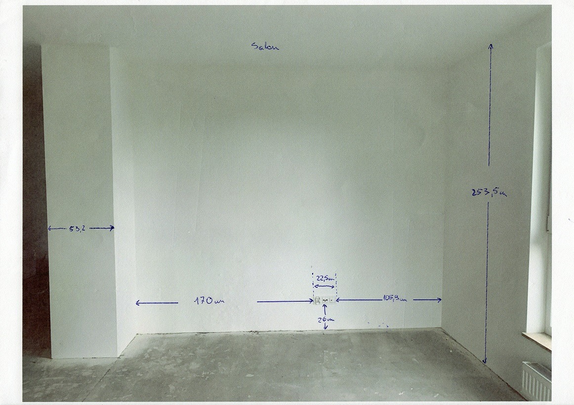 Pusty pokój w trakcie remontu z naniesionymi odręcznie wymiarami na ścianach, widoczne gniazdka elektryczne. Szary beton na podłodze, białe ściany. Salon w trakcie prac.