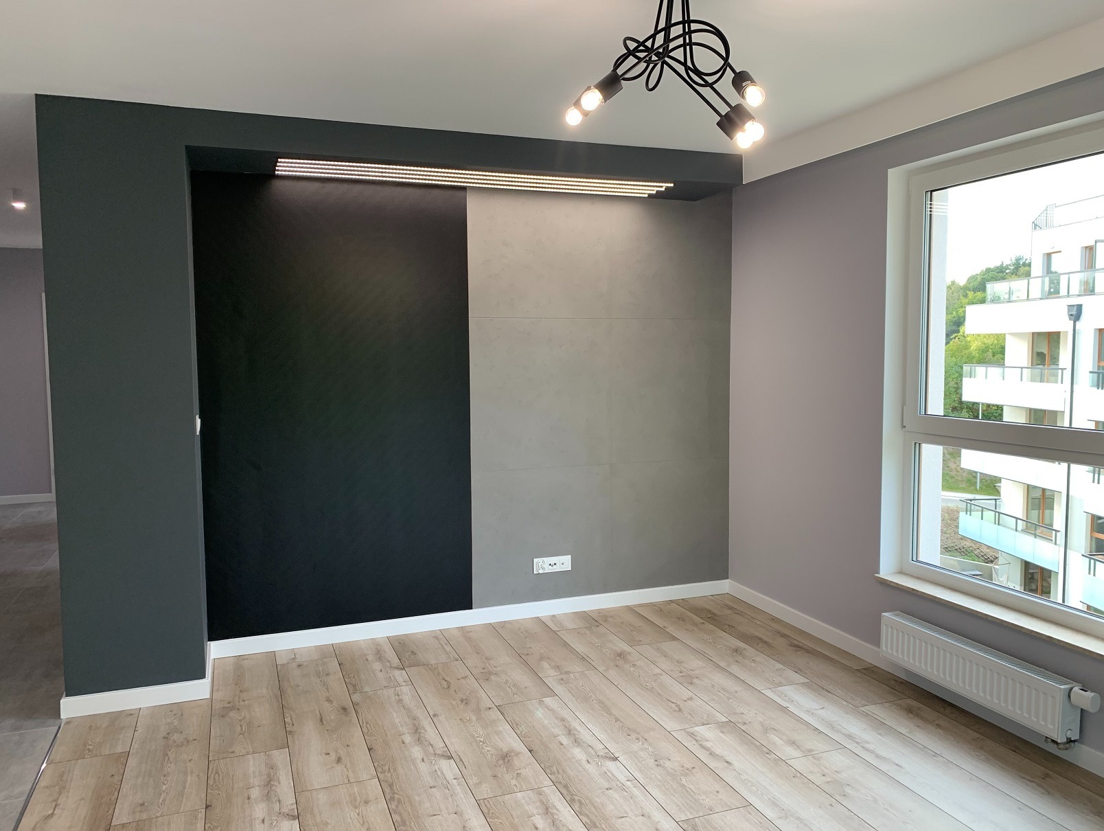 Nowoczesne wnętrze: drewnopodobne panele, ściana z szarą i czarną fakturą, designerska lampa sufitowa, okno z widokiem na zieleń. Minimalizm i elegancja.